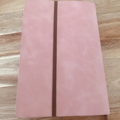 Caderno de capa rosa com elástico castanho sobre mesa de madeira clara