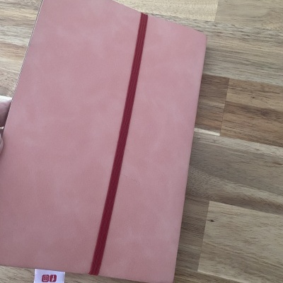 Caderno rosa com elástico vermelho e etiqueta numa superfície de madeira.