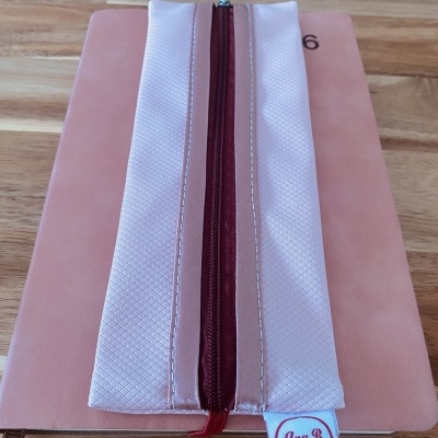 Estojo rosa claro com fecho vermelho e etiqueta Ana B Artesã sobre livro rosa