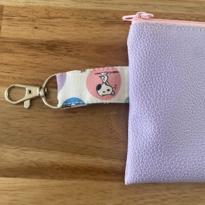 Carteira lilás com correia branca estampada com Snoopy num círculo cor-de-rosa e fecho rosa.