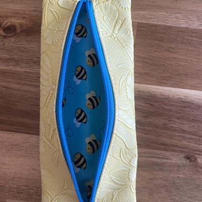 Estojo amarelo com fecho azul e interior azul com padrão de abelhas.