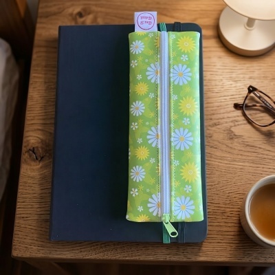 Estojo verde com padrão floral sobre capa de caderno preta numa mesa de madeira com óculos e chávena