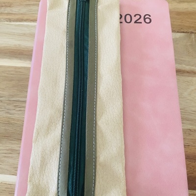 Estojo bege e verde com fecho de correr preto sobre caderno cor-de-rosa com número 2026