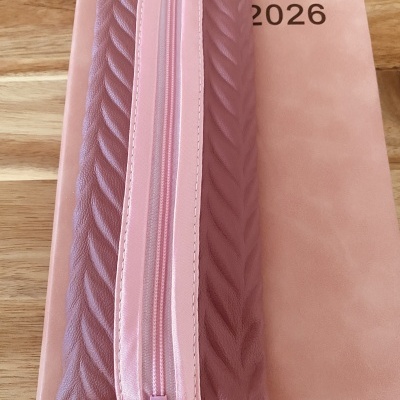 Estojo rosa com textura e fecho, sobre planner rosa com 2026, em mesa de madeira.