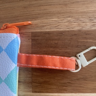 Bolsa pequena colorida com fecho laranja e mosquetão branco sobre mesa de madeira