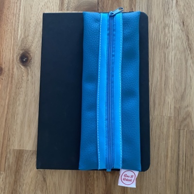 Estojo azul com fecho de correr azul e etiqueta Ana B Artesã sobre livro preto e madeira.
