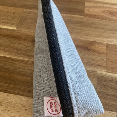 Estojo triangular cinzento claro com fecho preto e etiqueta branca com texto vermelho numa mesa de madeira.