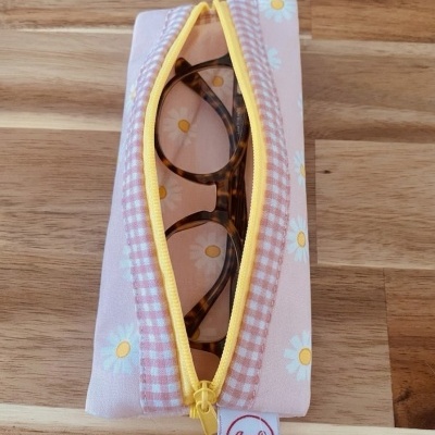 Estojo tecido rosa com margaridas e fecho amarelo, contendo óculos de armação castanha.
