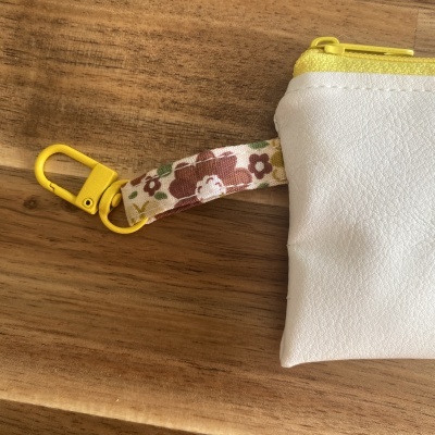 Bolsa pequena branca com fecho amarelo e cinta floral sobre superfície de madeira.