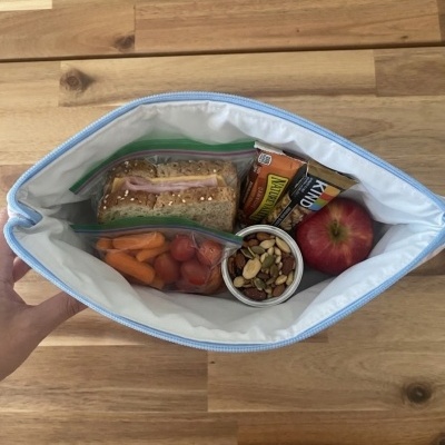Bolsa térmica aberta com lanche saudável em iços ziplock e recipiente, sobre mesa de madeira.