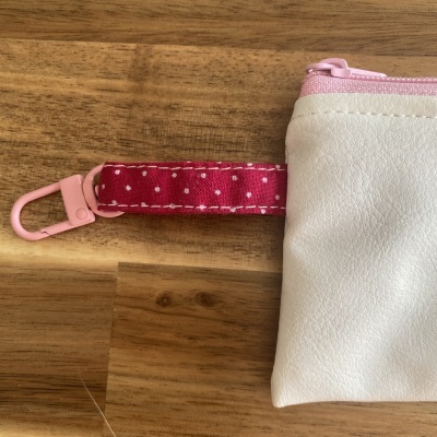 Bolsa pequena branca com fecho rosa e alça vermelha de bolinhas sobre madeira