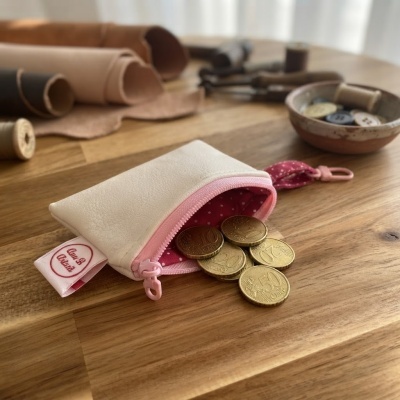 Carteira pequena branca com fecho rosa e moedas euros a sair, sobre mesa de madeira com rolos de couro e botões ao fundo