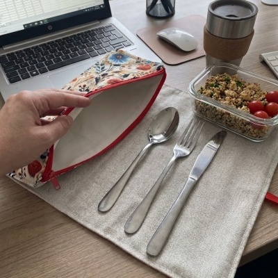 Mesa de escritório com computador portátil, talheres, nécessaire floral aberta, caixa com comida e caneca térmica.