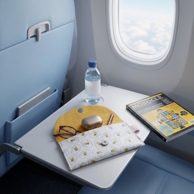 Estojo com padrão de abelhas, óculos, caixa metálica, caneta e revista MONOCLE em mesa de avião