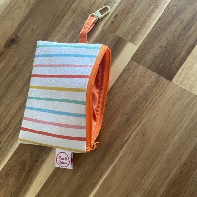 Bolsa colorida pequena com fecho laranja e etiqueta Ana B Artesã sobre madeira