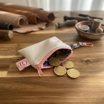Carteira pequena branca com fecho rosa e moedas euro à saída sobre mesa de madeira