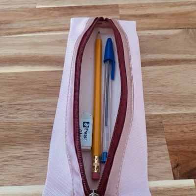 Estojo rosa aberto com lápis amarelo, caneta azul e borracha, sobre mesa de madeira clara.