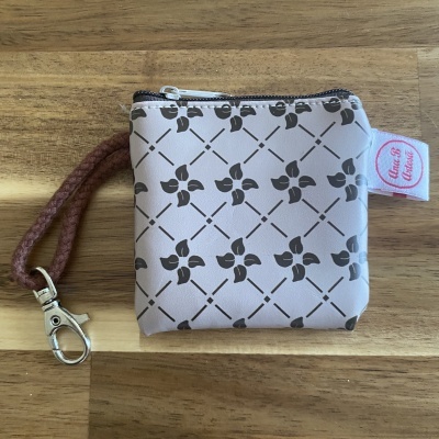 Bolsa pequena bege com padrão floral preto, zíper, cordão castanho e etiquetas em cima de superfície de madeira.