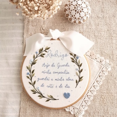 Bastidor bordado com frase, fita branca e decoração floral, sobre tecido bege com renda.