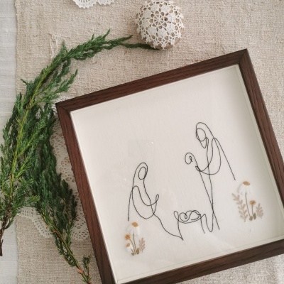 Quadro de madeira com ilustração minimalista e bordados, decorado com ramos verdes e crochê sobre tecido bege.