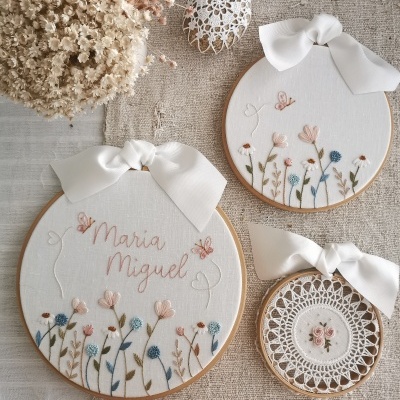 Três aros de bordado com flores e nome Maria Miguel, fitas brancas, sobre tecido cru com flores secas e ovo decorativo branco