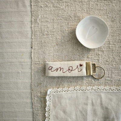 Porta-chaves de tecido bordado com texto e coração, pequeno prato branco e pano com renda sobre tecido bege texturado.