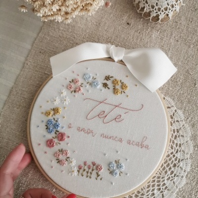 Bastidor redondo com bordado floral colorido e texto em vermelho sobre tecido branco, com laço branco e renda, visto numa mesa clara com flores secas ao lado.