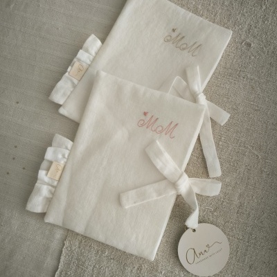 Dois cadernos tipo diário em tecido branco com bordados e fitas em superfície de tecido cinza/bege.