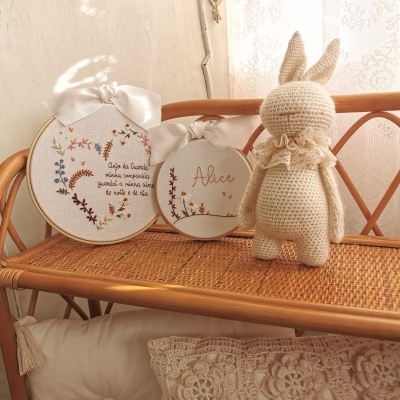 Dois bordados em aros de madeira com texto e um coelho de crochet sobre prateleira de rattan