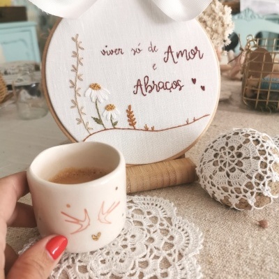 Bordado com texto motivacional acompanhado por chávena de café em ambiente com toalhas rendadas