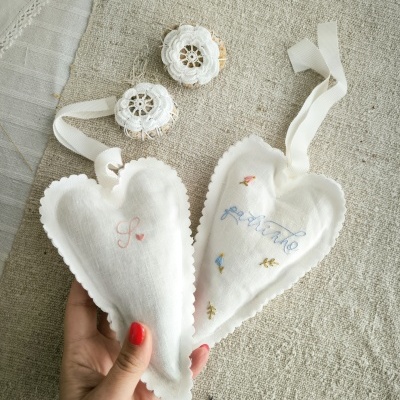 Dois corações de tecido branco com bordados e fita para pendurar sobre tecido bege.