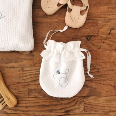 Saco branco de tecido com renda e cordão ao lado de sapatos de bebé em pele bege sobre superfície de madeira