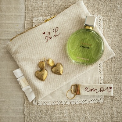 Bolsa de tecido bege com bordado AL, brincos dourados, perfume Chanel verde, chaveiro amor sobre tecidos neutros