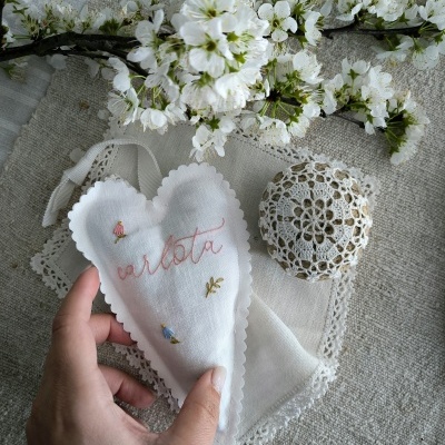Coração de tecido bordado com nome Carlota segurado à mão ao lado de ovo com renda e flores brancas