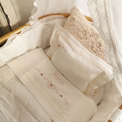 Berço de bebé com roupa de cama branca e bege bordada com nome 'Maria' e almofada rendada.