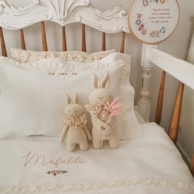 Cama branca com almofadas bordadas, coelhos de croché e bastidor bordado com texto em português