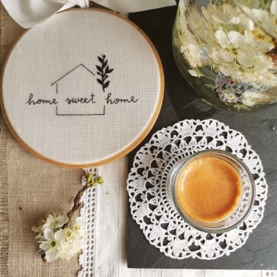 Bastidor bordado com frase home sweet home, chávena de café num pires de crochet, jarro com flores e tecido de linho