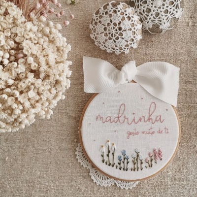 Bordado em bastidor de madeira com frase para madrinha, laço branco e flores bordadas, ao lado de flores secas e bolas de croché