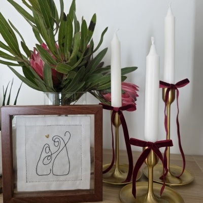 Castiçais dourados com velas brancas e laços vermelhos, moldura de madeira com bordado minimalista e vaso com flores e folhas verdes