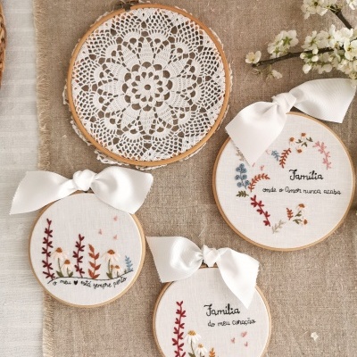 Quatro bordados circulares com fita branca e padrão floral, incluindo frases em português, sobre tecido bege cinzento e flores brancas ao lado.