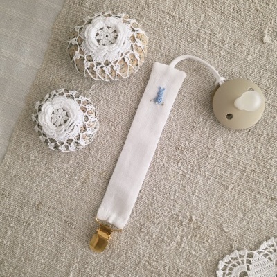 Presilha com tecido branco e bordado azul pendurada em chupeta branca sobre tecido bege com crochê.