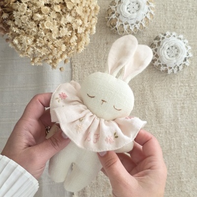 Brinquedo de pelúcia coelho branco segurado por mãos, com babador rosa bordado
