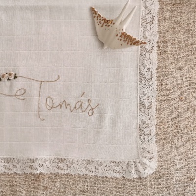 Toalha branca com bordado nome Tomás e renda, concha ao lado sobre tecido bege texturizado