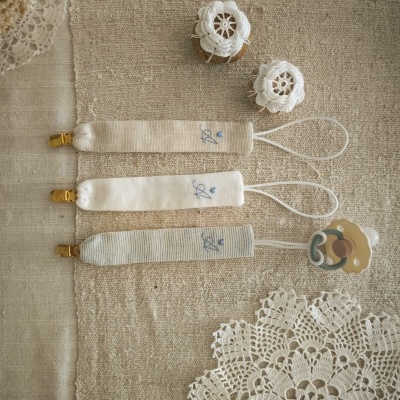 Três pegas para chupetas claras com chupetas em fundo bege e toalha de crochê branca