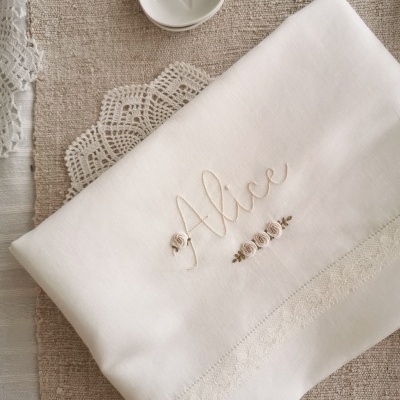 Toalha branca bordada com nome Alice e flores, com prato branco e renda em fundo bege.