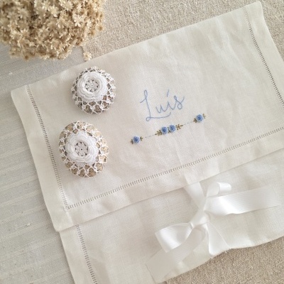 Toalha de linho branca bordada com nome Luís e flores azuis, com fita branca e sacos de crochê branco com pedras em tecido bege.