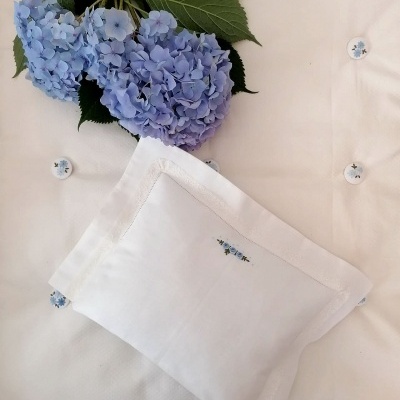 Jogo de cama branco com botões e bordados de flores azuis e ramo verde, com flores hortênsia azuis ao lado