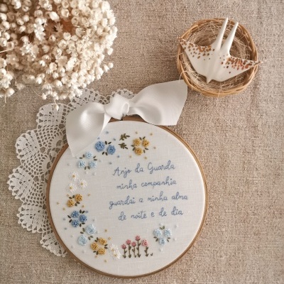 Bordado artesanal com texto e flores em bastidor de madeira sobre tecido bege com flores secas e figura de pássaro decorativo