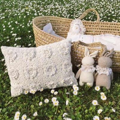 Berço de verga com roupa branca, almofada de crochê branca e dois bonecos de coelho em crochê em relva com flores brancas