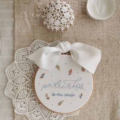 Bastidor bordado com frase e flores, laço branco, sousplat rendado e recipiente branco sobre tecido bege.